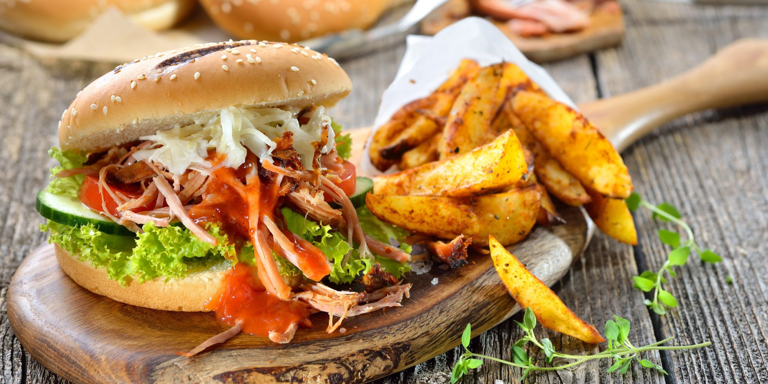 Sandwich aus Sesambrötchen mit Salat und pulled pork, dazu knusprige Kartoffelspalten in der Papiertüte – Barbecue pulled pork sandwich with coleslaw, hot BBQ sauce and potato wedges in a paper bag