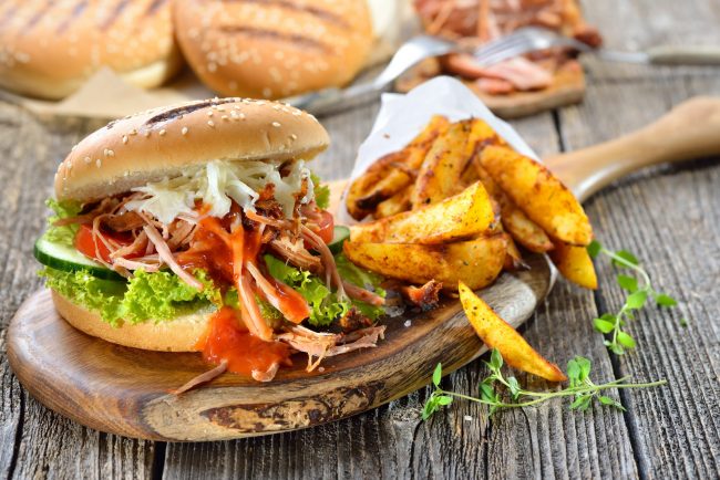 Sandwich aus Sesambrötchen mit Salat und pulled pork, dazu knusprige Kartoffelspalten in der Papiertüte – Barbecue pulled pork sandwich with coleslaw, hot BBQ sauce and potato wedges in a paper bag